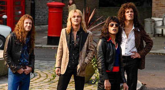 Four men with long hair dressed in 1970s rock style standing on a city street.
