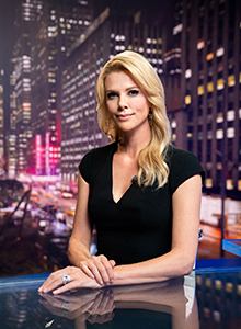 A blonde woman in a black V-neck dress poses against a nighttime cityscape background.
