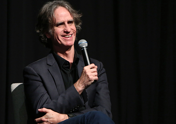 A smiling man in a dark suit holds a microphone while seated against a black background.