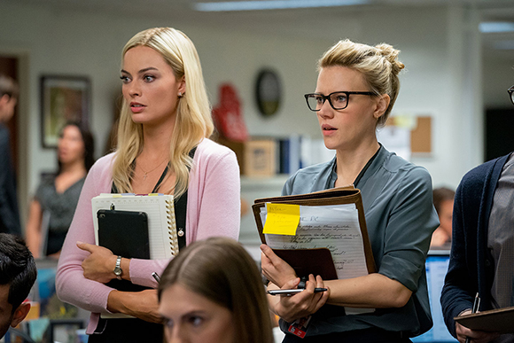 Two women in an office setting, one blonde in a pink cardigan and one blonde with glasses in a grey shirt.