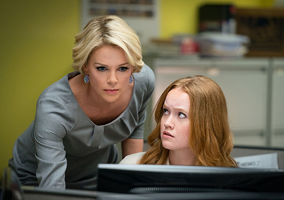 A blonde woman in a grey dress leans over a redhead looking up at her in an office setting.
