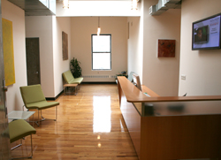 A bright office lobby with a long wooden reception desk, polished wood floors, and green chairs.