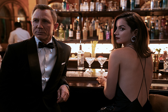 A man in a tuxedo and a woman in a backless black dress sit at a dimly lit bar with bottles in the background.