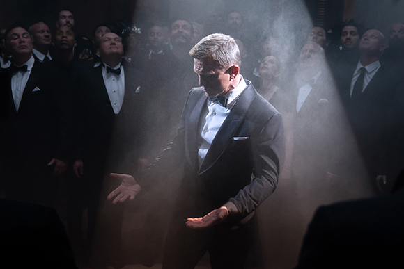 A man in a tuxedo stands in a spotlight amidst a crowd of men in formal attire and hazy smoke.