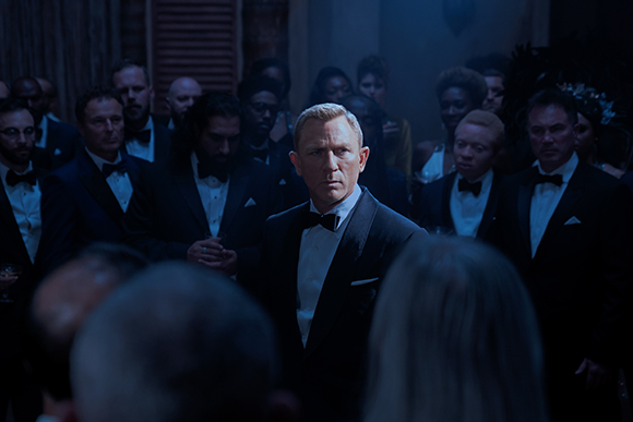A man in a black tuxedo and bowtie looks forward amidst a crowd of people in formal wear in a dimly lit room.