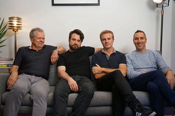 Four men of varying ages sitting together on a grey sofa in a brightly lit room.