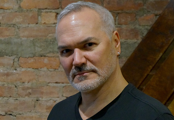 A man with short gray hair and a gray beard looking toward the camera against a brick wall.