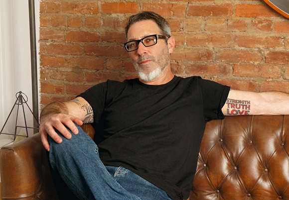 A man with glasses and a grey beard wearing a black t-shirt, sitting on a brown leather sofa against a brick wall.