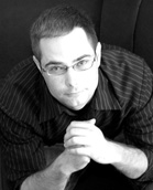 Black and white portrait of a man with short hair and glasses wearing a dark striped shirt.