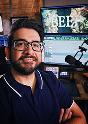 A man with glasses and a beard smiling in front of computer monitors in a studio.