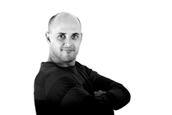 A bald man in a dark long-sleeved shirt stands with his arms crossed against a white background.
