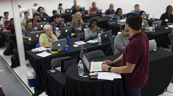 A man in a maroon shirt stands at the front of a classroom facing students seated at black tables with laptops.