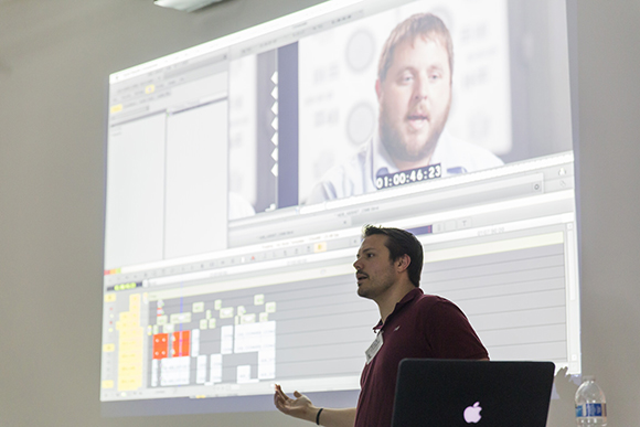A man in a maroon shirt gestures while presenting a video editing software interface on a large screen.