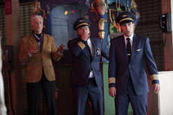 Two men in navy pilot uniforms and one man in a tan blazer stand in a dimly lit room with surprised expressions.