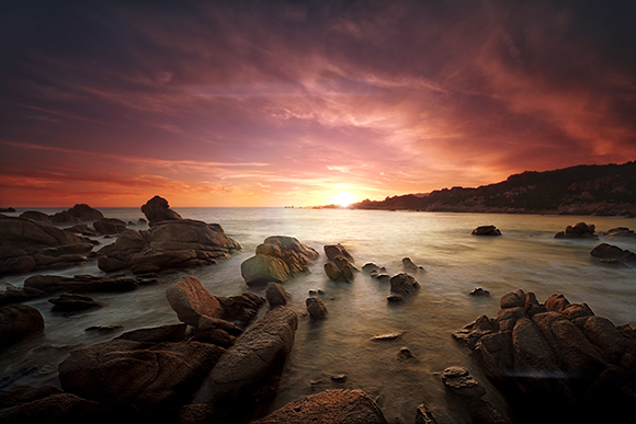 A vibrant sunset glows over a rocky ocean shore with waves washing around large stones.