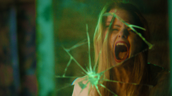 A woman with blonde hair screams through a pane of shattered, spiderwebbed glass.