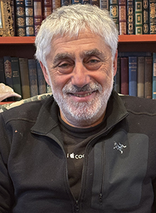 A smiling man with grey hair and a beard wearing a black jacket in front of a bookshelf.
