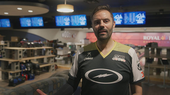 A man wearing a black and white Storm brand jersey stands in a bowling alley with monitors in the background.
