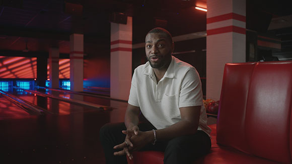 A man in a white polo shirt sits on a red booth inside a dimly lit bowling alley.
