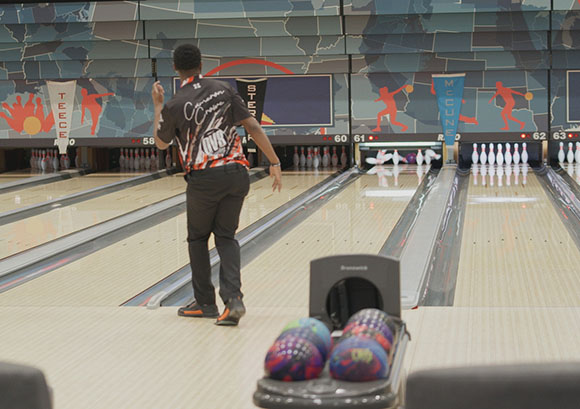 A man in a black and orange jersey stands on a bowling lane, facing the pins.