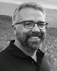 Black and white portrait of a smiling man with glasses and a beard outdoors.