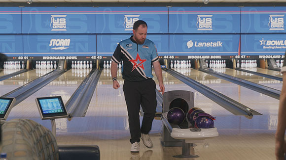 A man in a blue and black polo shirt walks past bowling balls on a rack in a bowling alley.