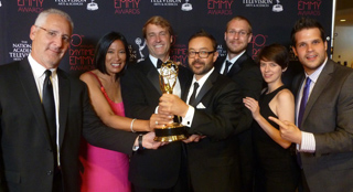 A group of people in formal attire smiling and holding a gold Emmy award trophy.