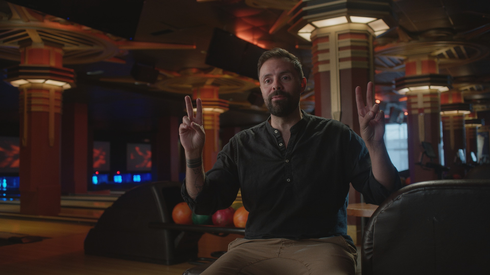 A bearded man in a dark shirt sits in a bowling alley, holding up two fingers in peace signs on both hands.