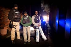 Three men wearing tactical vests and gear stand together in a dark alleyway at night.