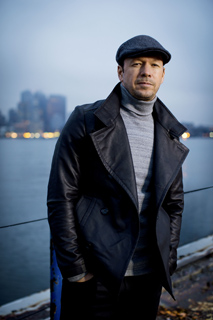 A man wearing a black leather jacket, grey turtleneck, and a dark flat cap standing outdoors.