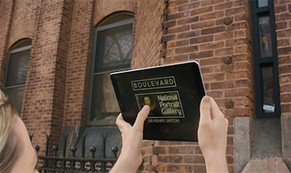 A person holds a tablet displaying a National Portrait Gallery screen in front of a brick building.