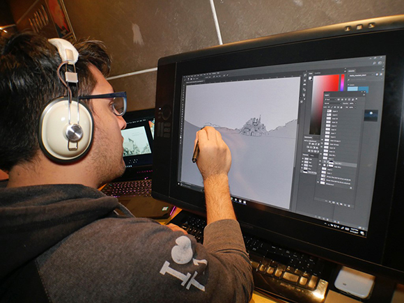 A person wearing headphones uses a stylus to draw a landscape on a large computer monitor.