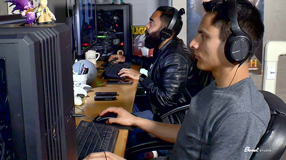 Two men wearing headphones sit at desks using computers and mice in a dimly lit room.