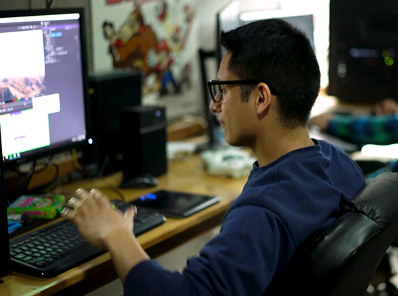 A man with glasses sits at a desk using a computer with a monitor, keyboard, and speakers.