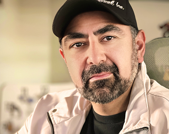 Close-up portrait of a man with dark hair, a salt-and-pepper beard, and a black baseball cap.