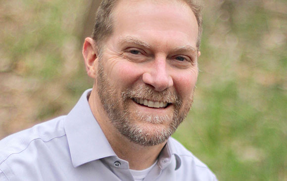 A smiling man with short brown hair and a salt-and-pepper beard wearing a light purple button-down shirt.