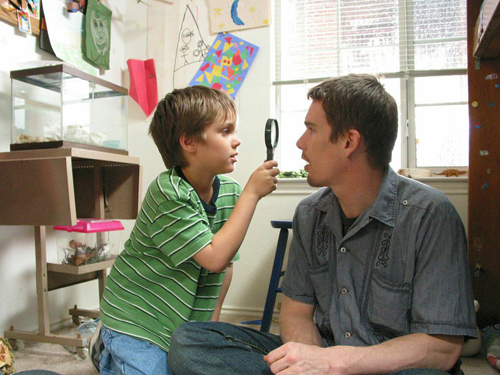 A young boy in a green striped shirt holds a magnifying glass up to a man's face.