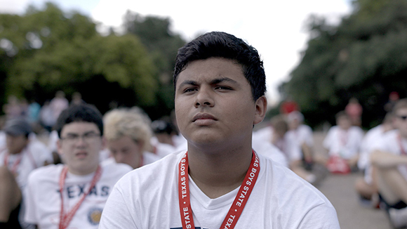 A young man in a white t-shirt and red lanyard looks forward with a serious expression outdoors.