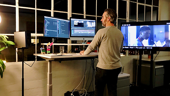 A man stands at a desk with three computer monitors displaying video editing software and footage.
