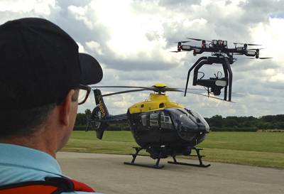 A man in a black cap watches a large black drone hovering near a black helicopter on a runway.