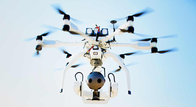 A white six-rotor drone with a spherical camera gimbal flying in a clear blue sky.