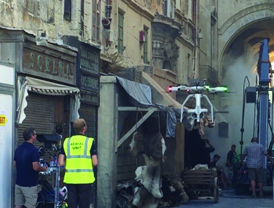 A man in a neon yellow vest stands near a white drone flying in front of a building set.
