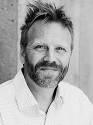 Black and white headshot of a man with short, messy hair and a salt-and-pepper beard smiling slightly.