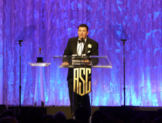A man in a black tuxedo and bowtie speaks behind a clear podium with a gold ASC logo against a blue backdrop.
