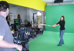 A woman in a black shirt and jeans holds a prop sword in front of a large green screen.