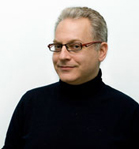 A man with graying hair and glasses wearing a black turtleneck against a white background