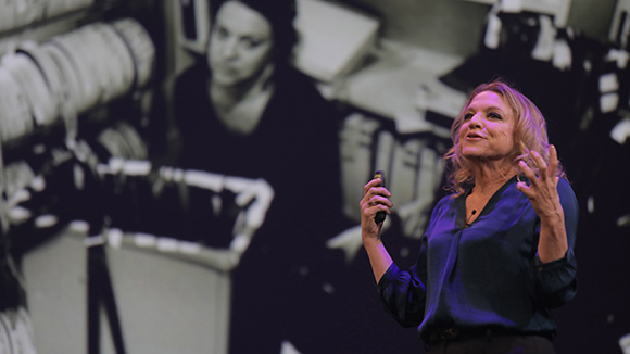 A woman in a blue blouse gestures with her hands while speaking on a stage.