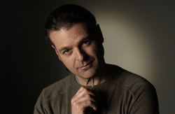 A man with dark hair wearing a brown sweater looks toward the camera against a dark background.