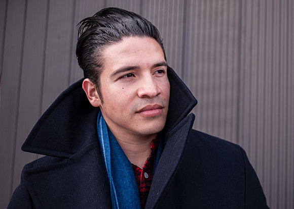 A man with dark hair wearing a black coat and blue scarf looks off to the side against a gray ribbed wall.