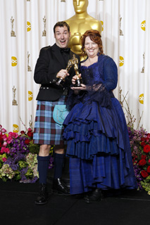 A man in a kilt and a woman in a blue gown hold an Oscar statuette against a gold backdrop.